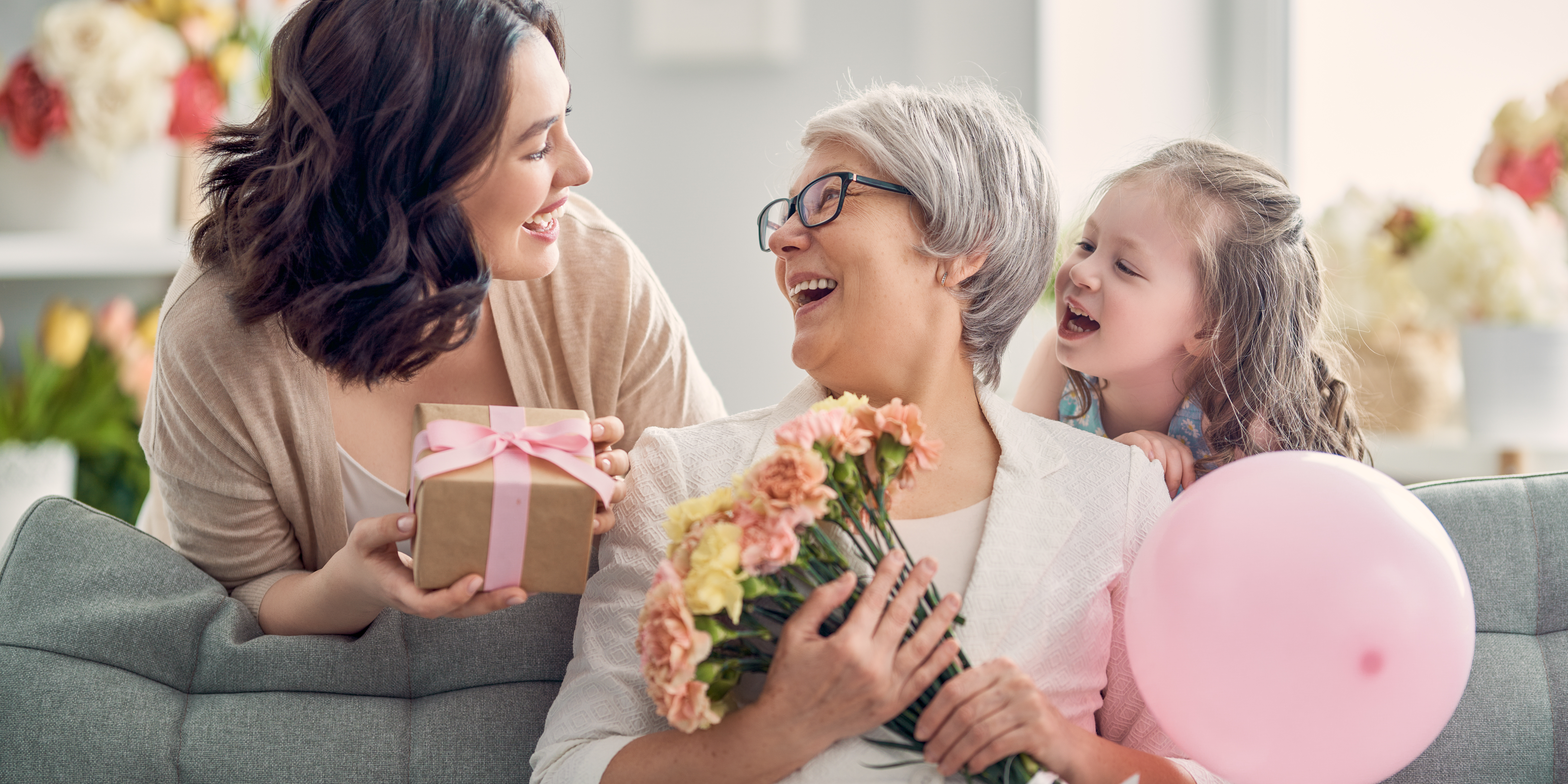 Three generations of women — a grandmother, mother, and young daughter — sharing gifts, flowers, and laughter together on Mother’s Day.