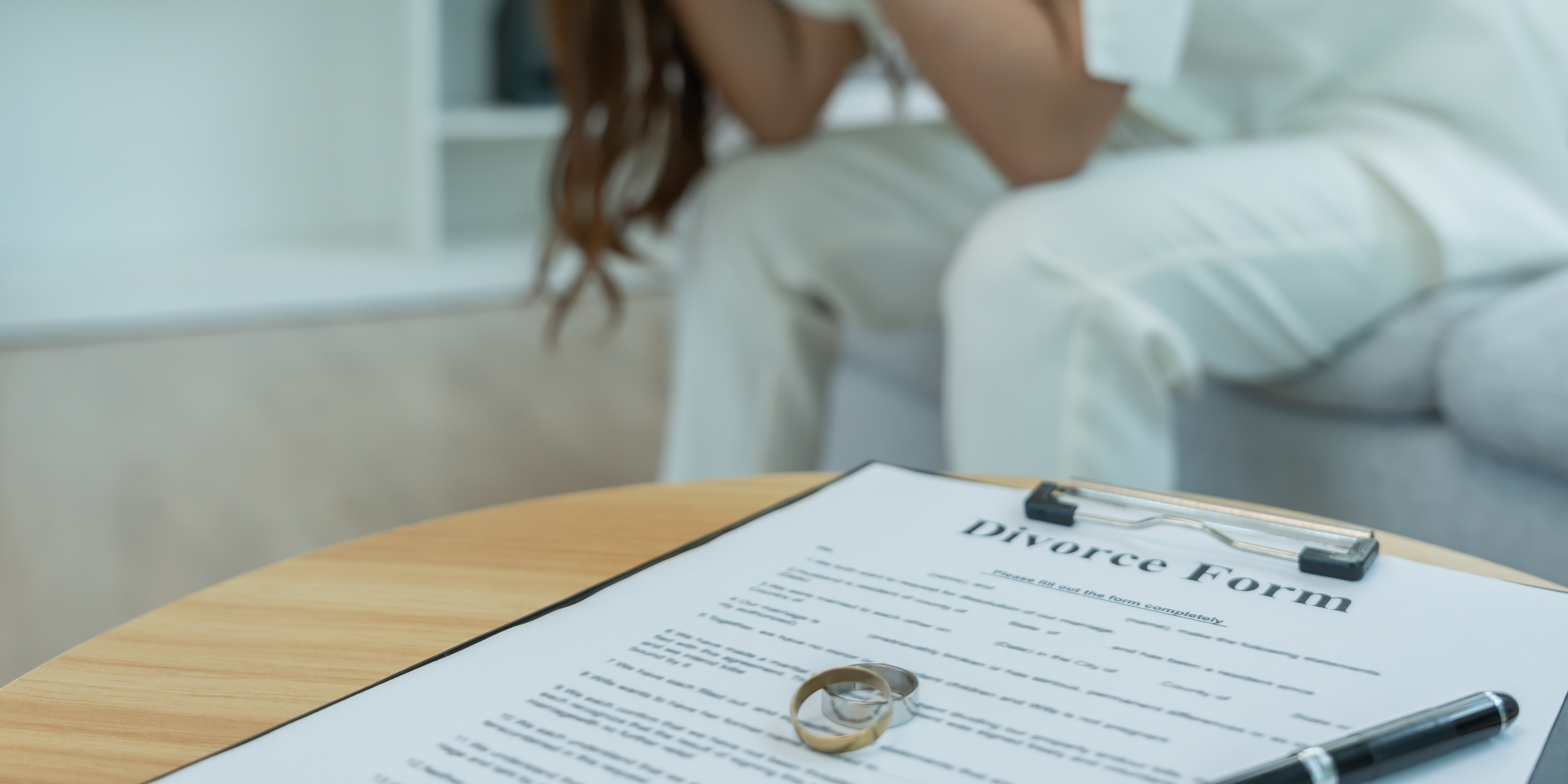 A person sitting with their head in their hands in the background, with a divorce form, wedding rings, and a pen on a table in the foreground.