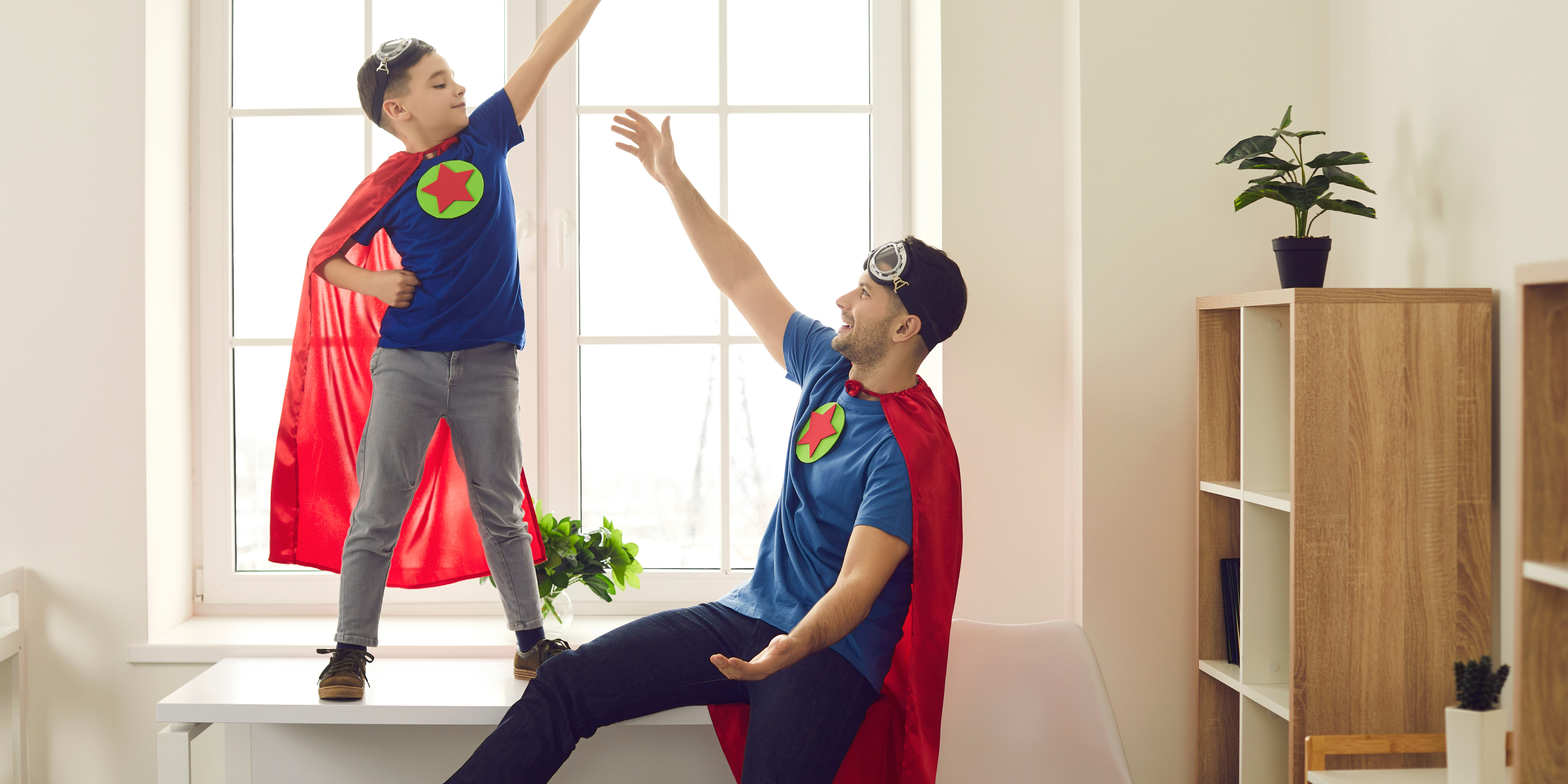 A father and young son dressed as superheroes, wearing matching capes and shirts with star emblems, playing together in a bright room.