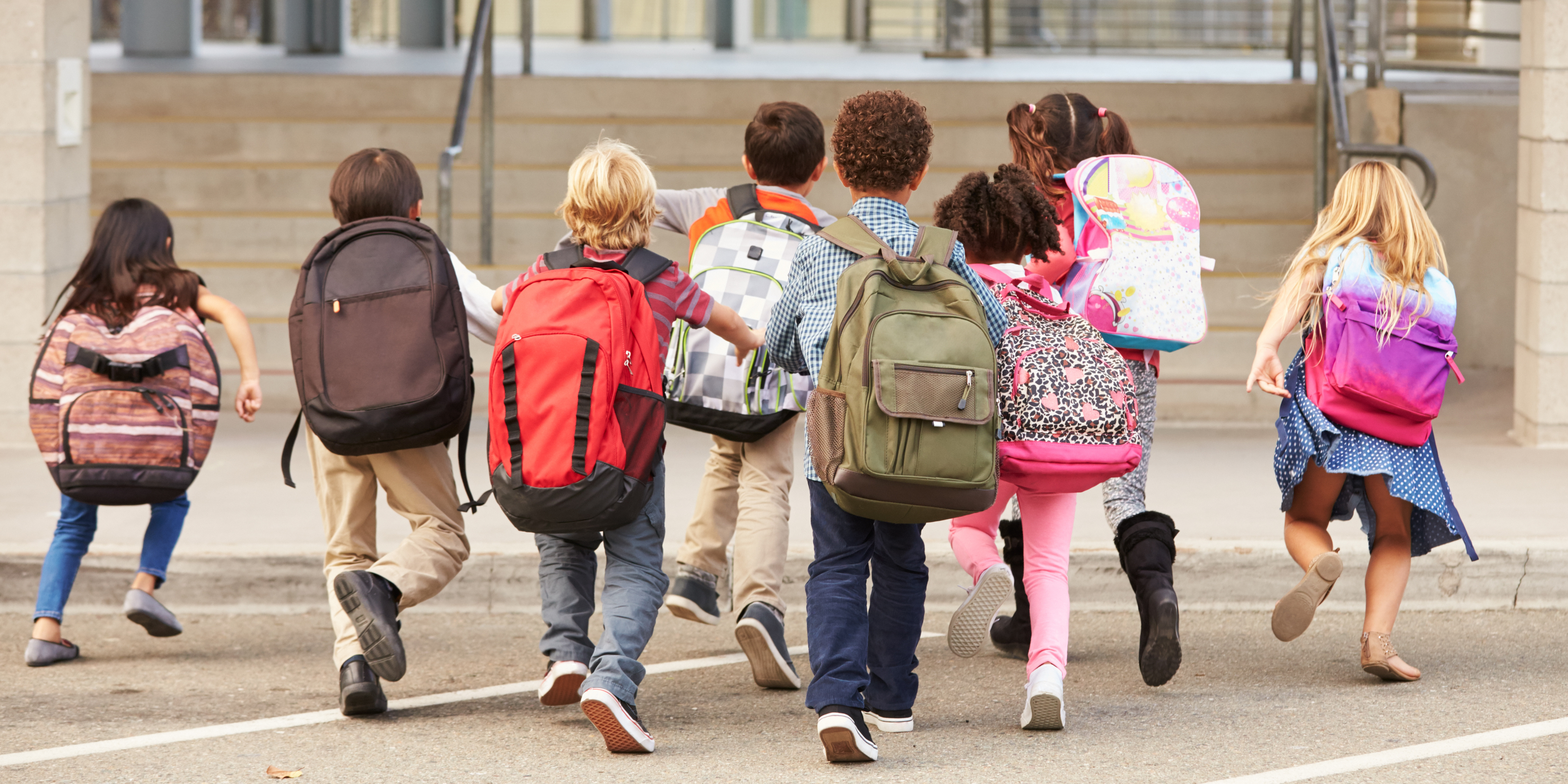 A group of young children wearing backpacks running toward a school entrance, viewed from behind.