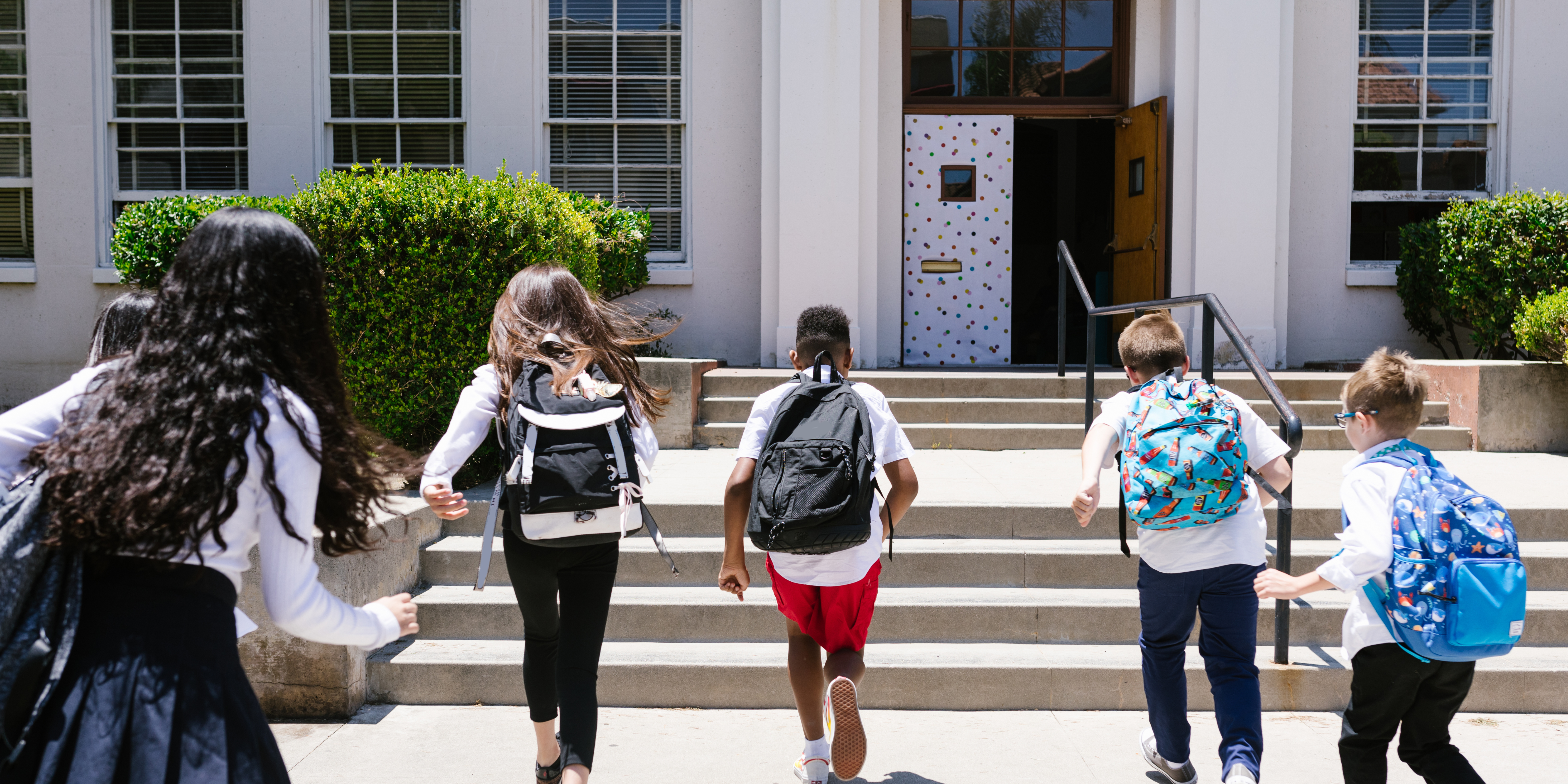Group of children wearing backpacks running toward a school building, symbolizing back-to-school season and co-parenting coordination after divorce.