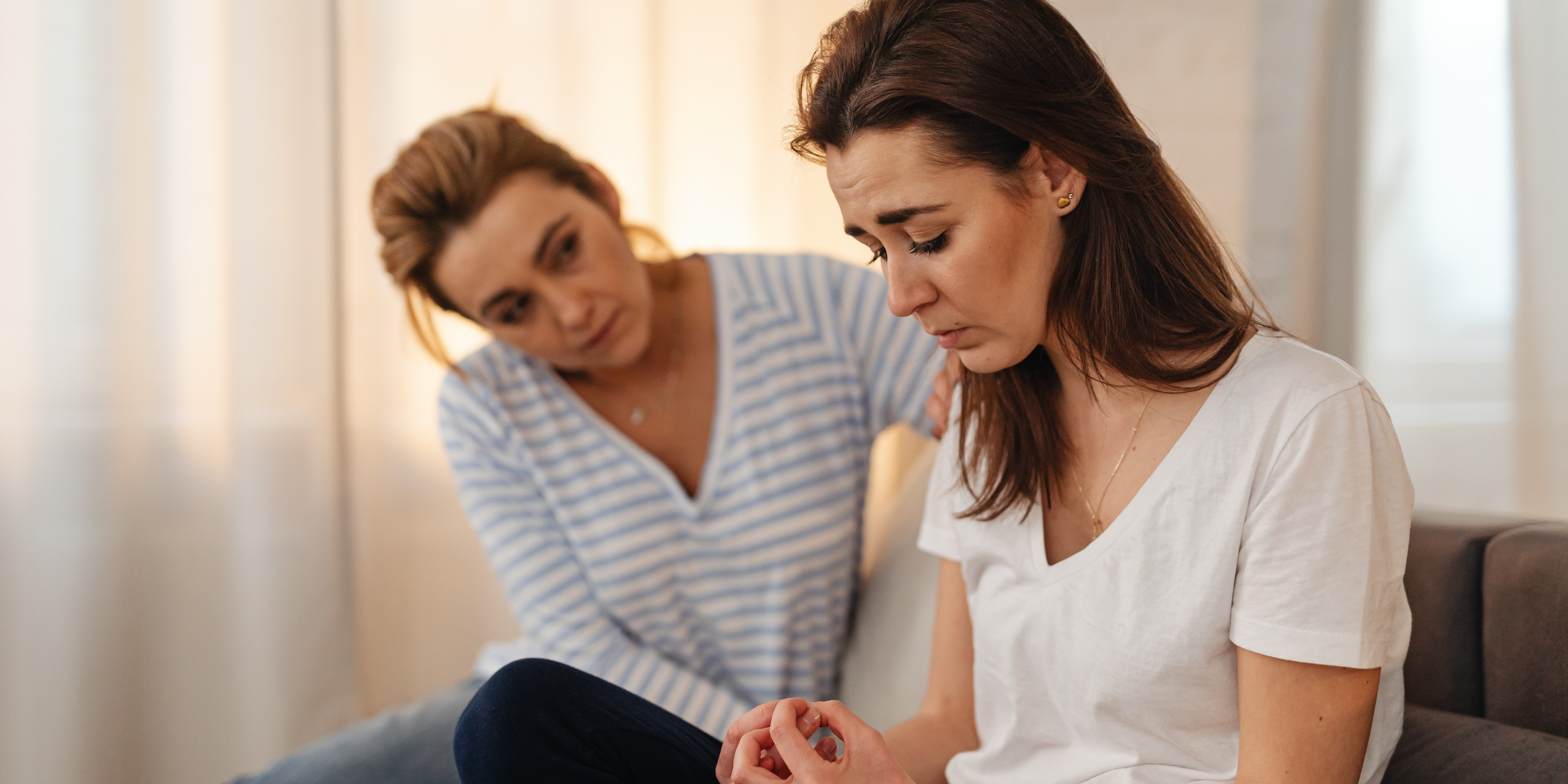 A woman sitting beside a sad friend, offering comfort with a hand on her shoulder.