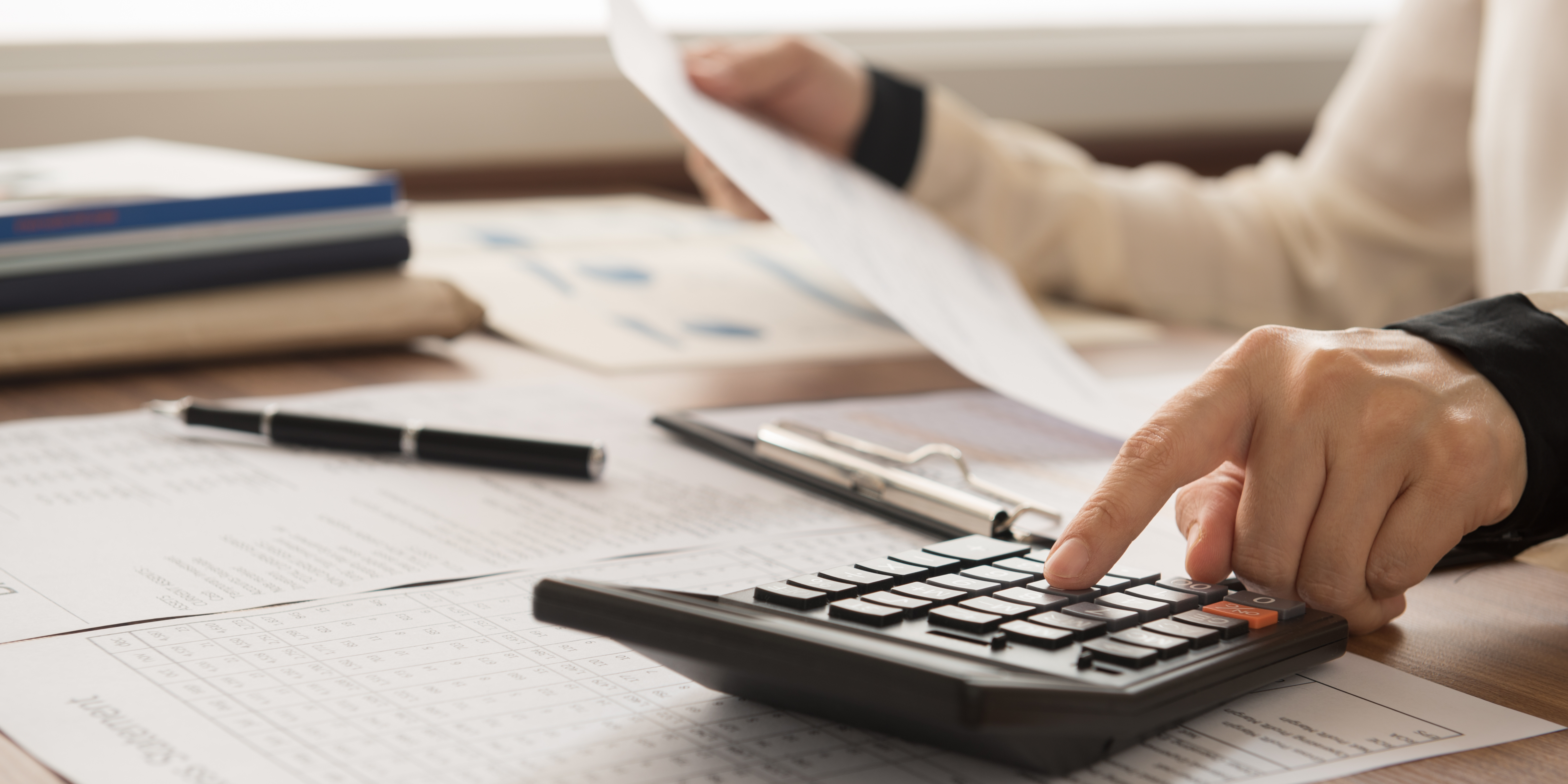 A person reviewing financial documents at a desk, using a calculator while holding printed paperwork.