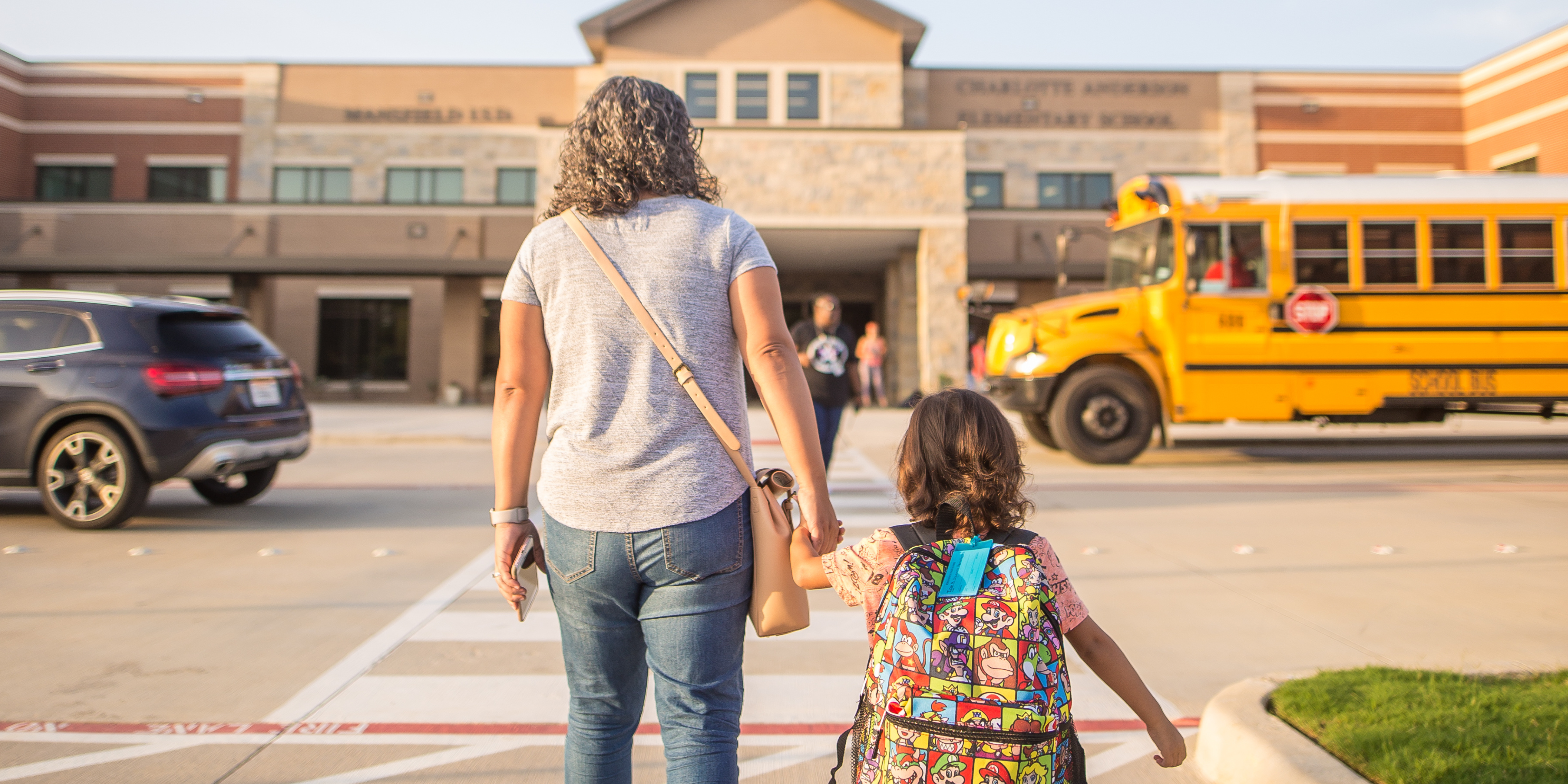 A parent walks hand in hand with their young child toward an elementary school as a yellow school bus passes nearby.