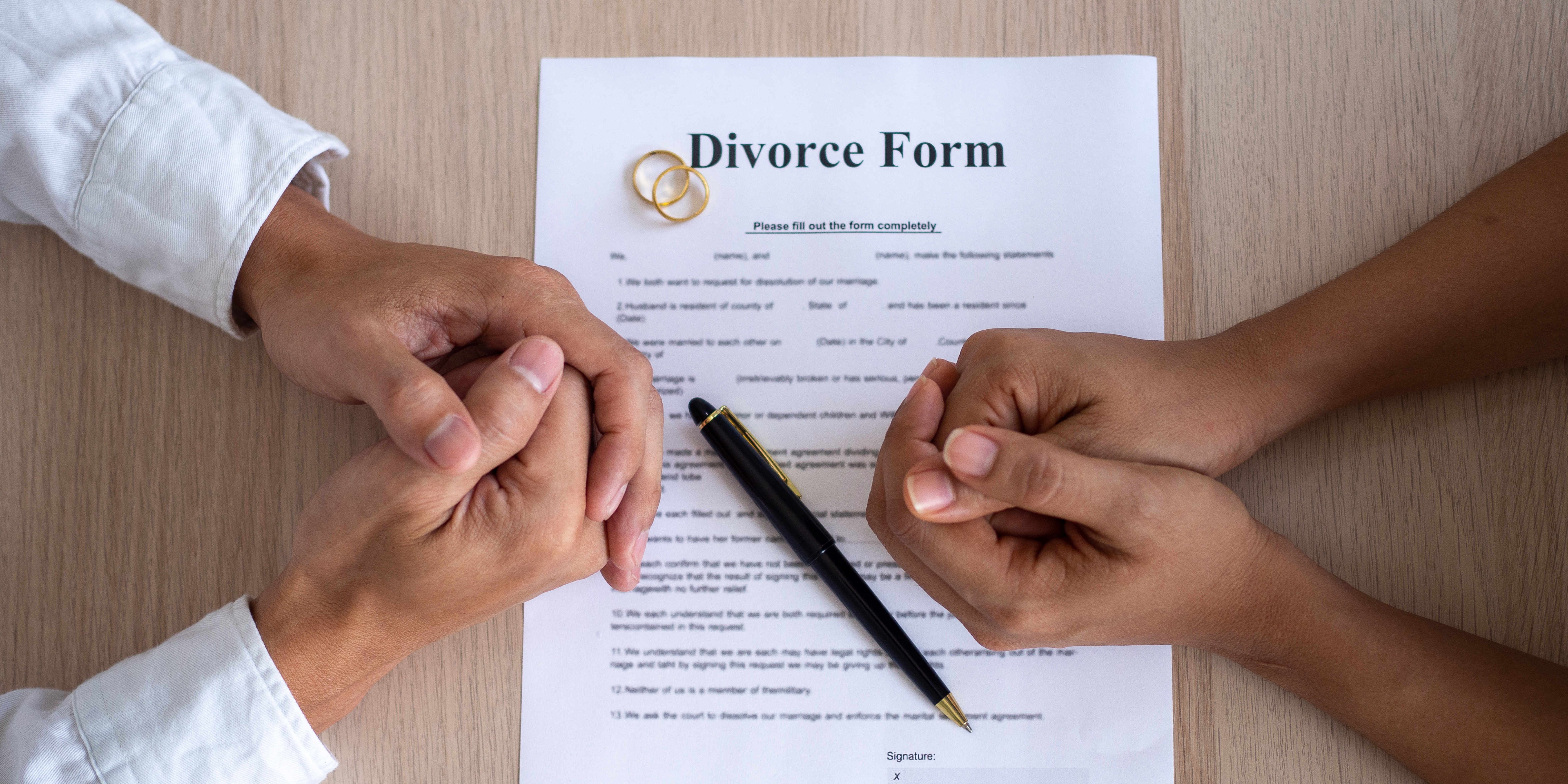 A close-up of two people sitting across a divorce form on a table, hands clasped tensely, with wedding rings and a pen placed on top of the paperwork.