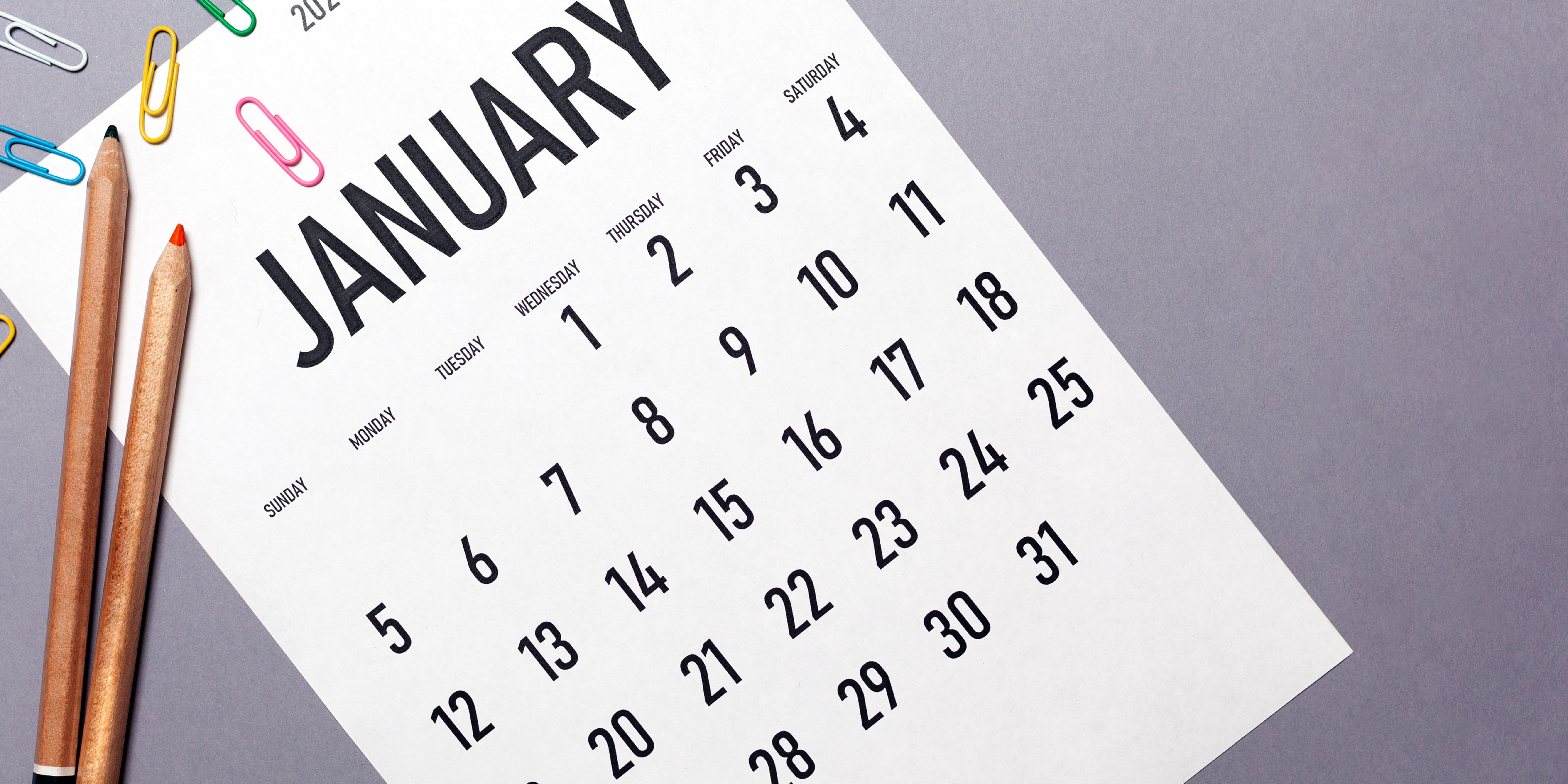 A paper calendar opened to the month of January, surrounded by pencils and colorful paperclips.
