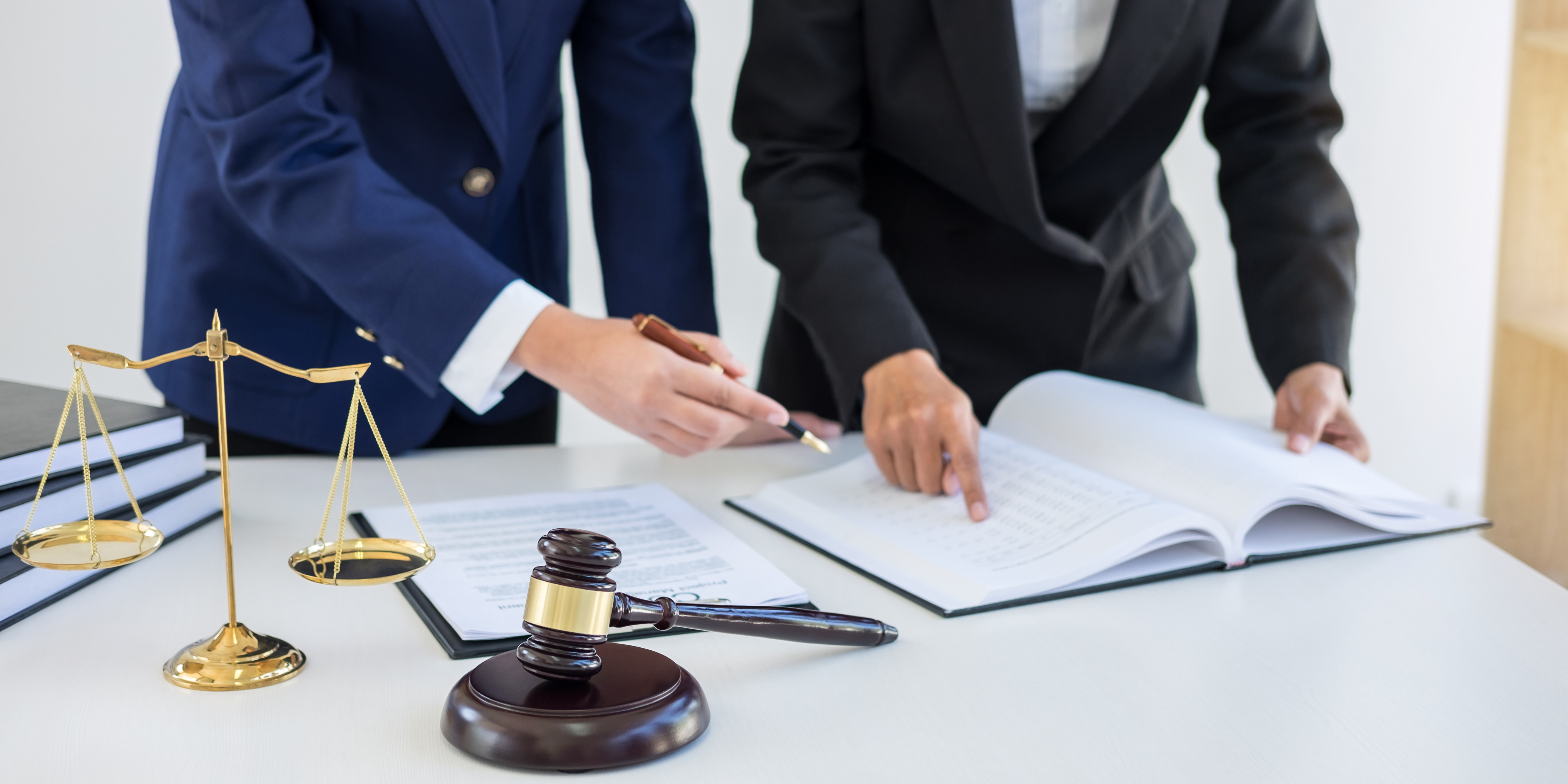 Two professionals in business attire reviewing legal documents at a desk with a gavel and scales of justice nearby.