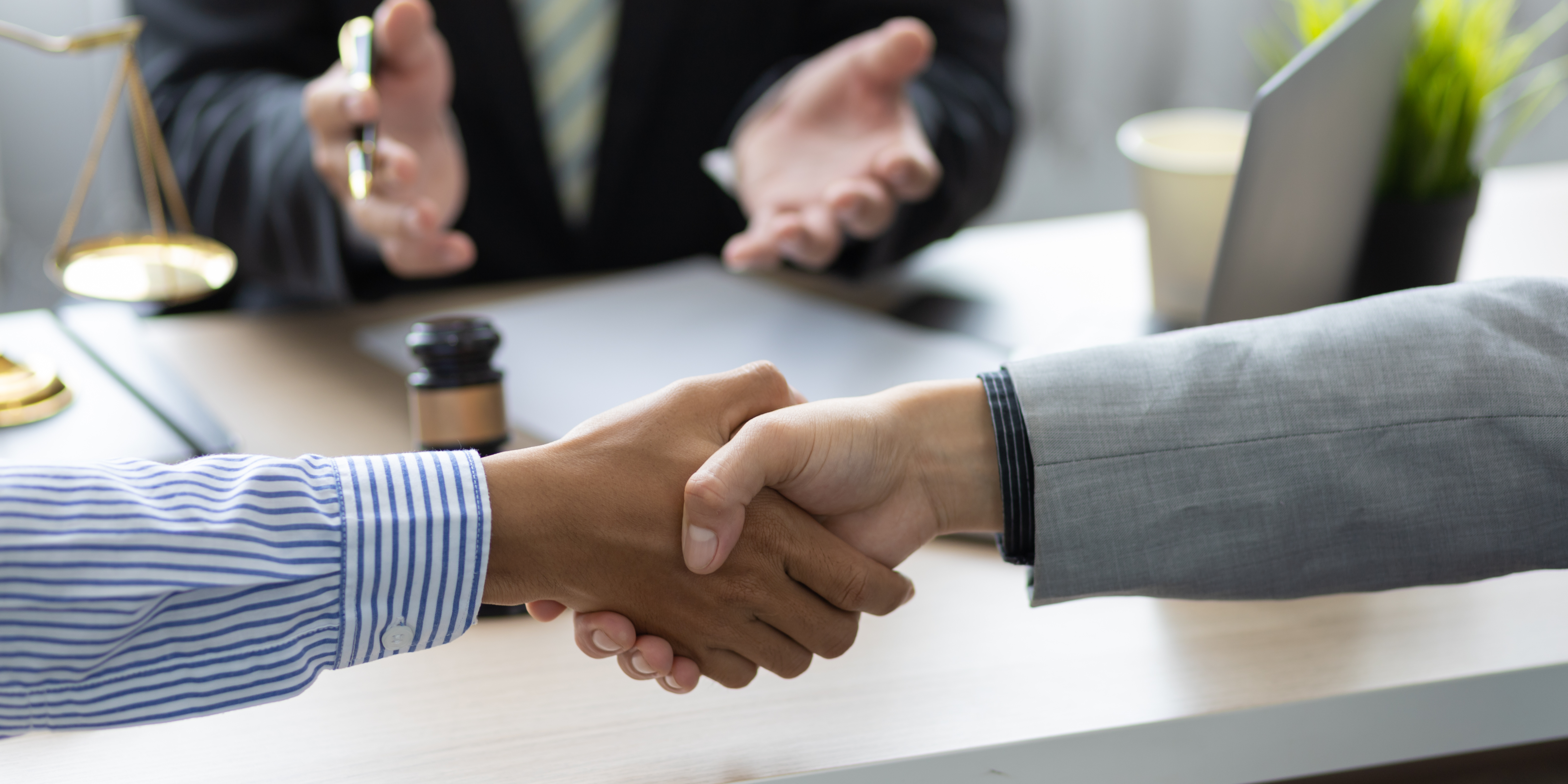 Two people shaking hands at a desk while a mediator in professional attire gestures and guides the discussion, with legal documents and scales of justice nearby.