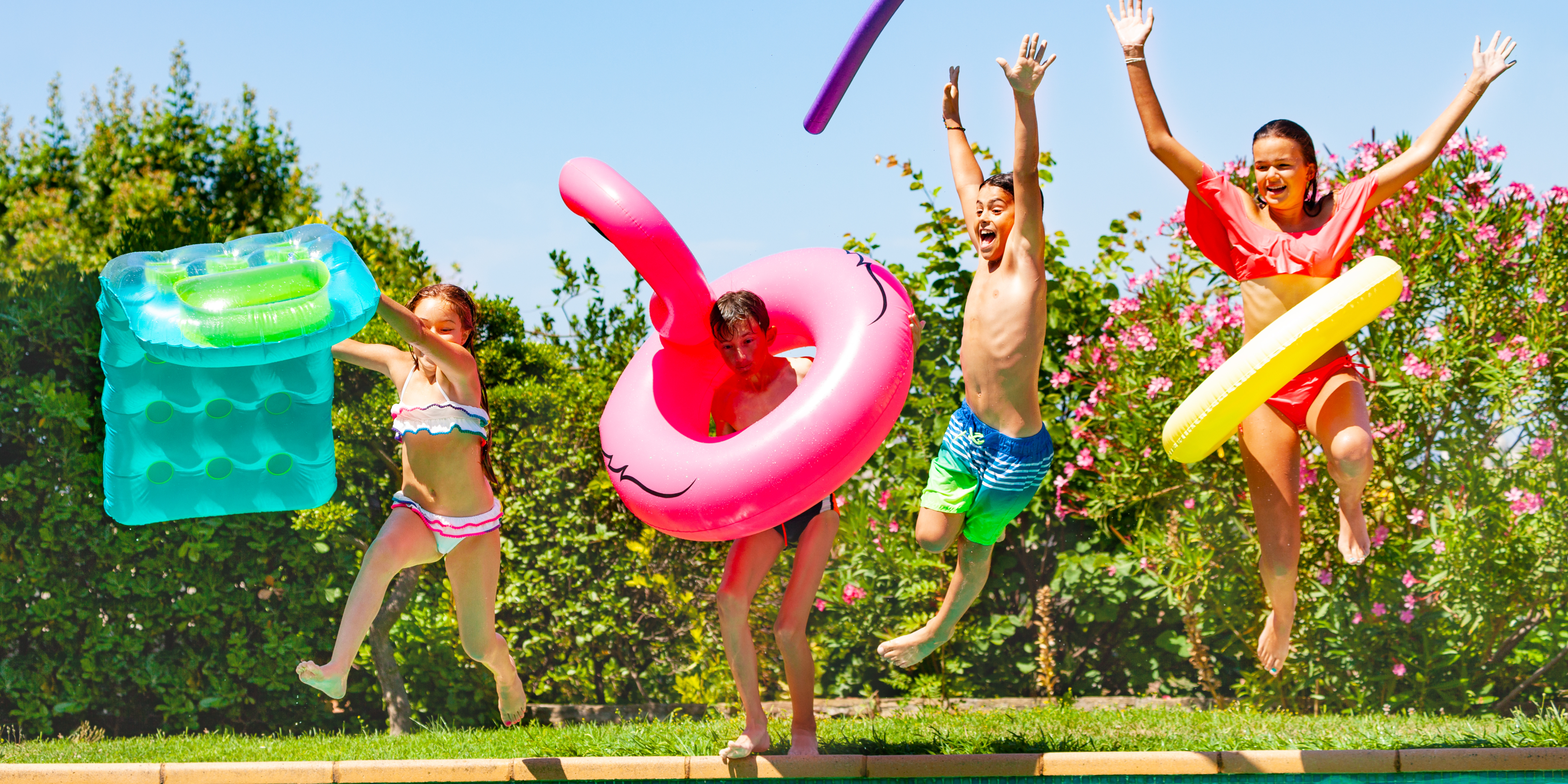 Four children jump into a swimming pool on a sunny day, holding colorful pool floats and smiling.
