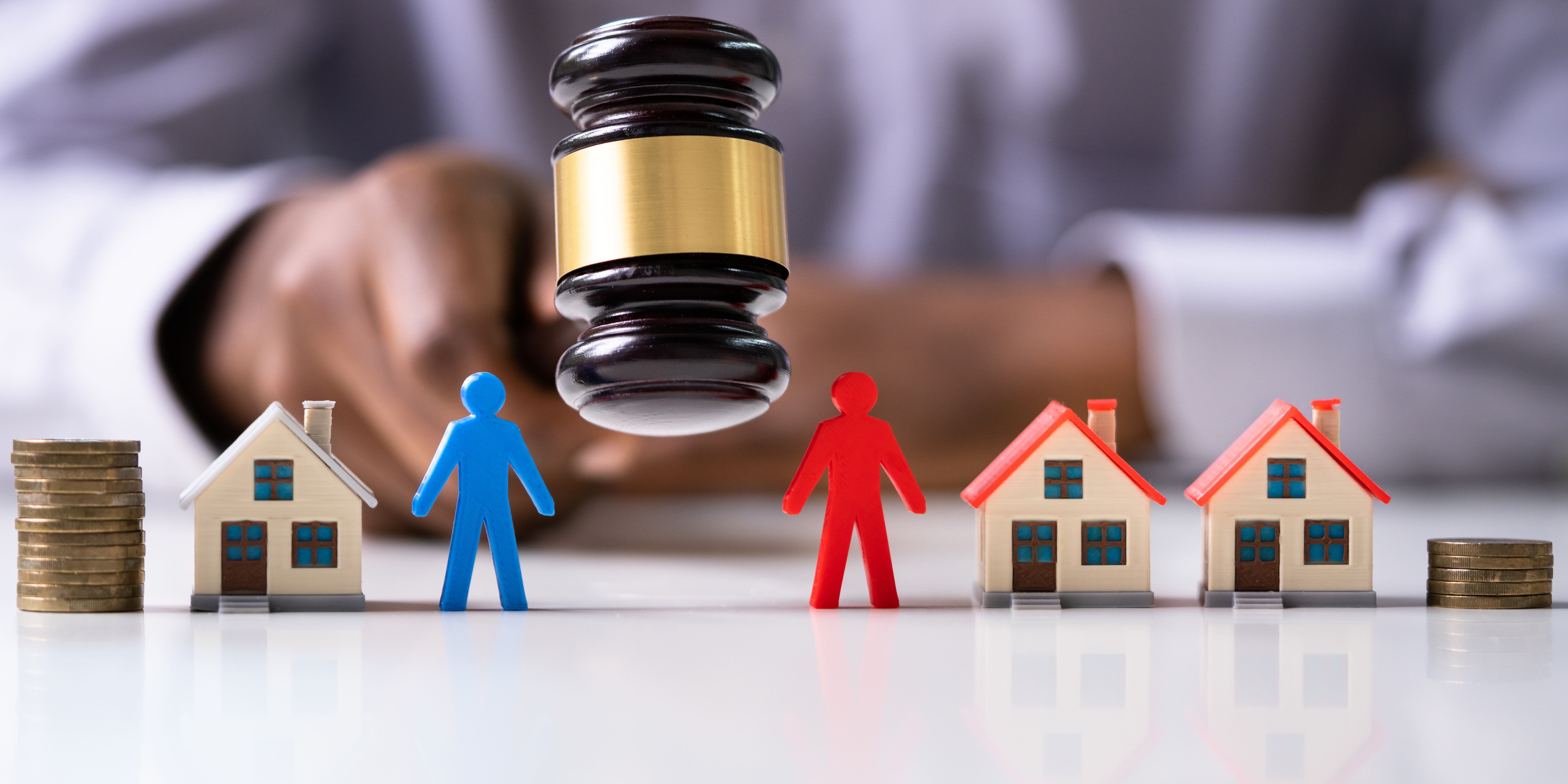 A judge’s gavel over miniature houses, coins, and colored human figurines representing spouses during asset division.