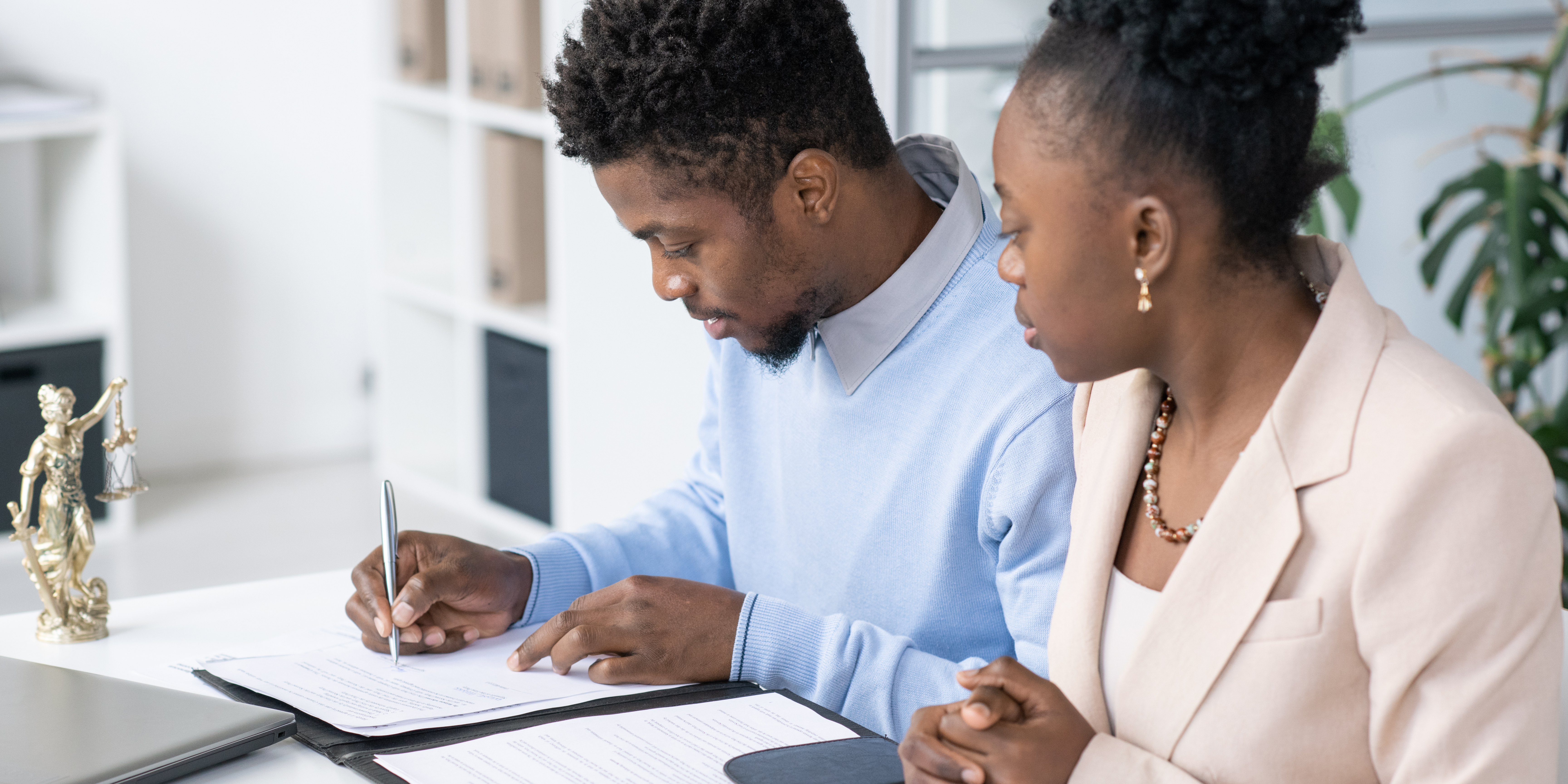 Couple reviewing and signing legal documents at a desk beside a Lady Justice statue, symbolizing cooperation, understanding, and mutual agreement in an uncontested divorce.